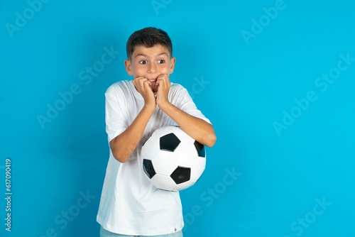 Fearful Little hispanic boy wearing white T-shirt holding a football ball keeps hands near mouth, feels frightened and scared,  has a phobia,  Shock and frighted concept. © Jihan