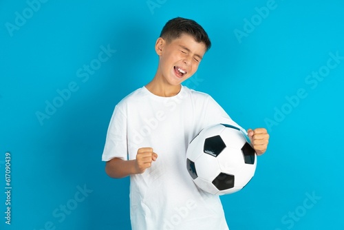 Ecstatic Little hispanic boy wearing white T-shirt holding a football ball shout loud yeah fist up raise win lottery