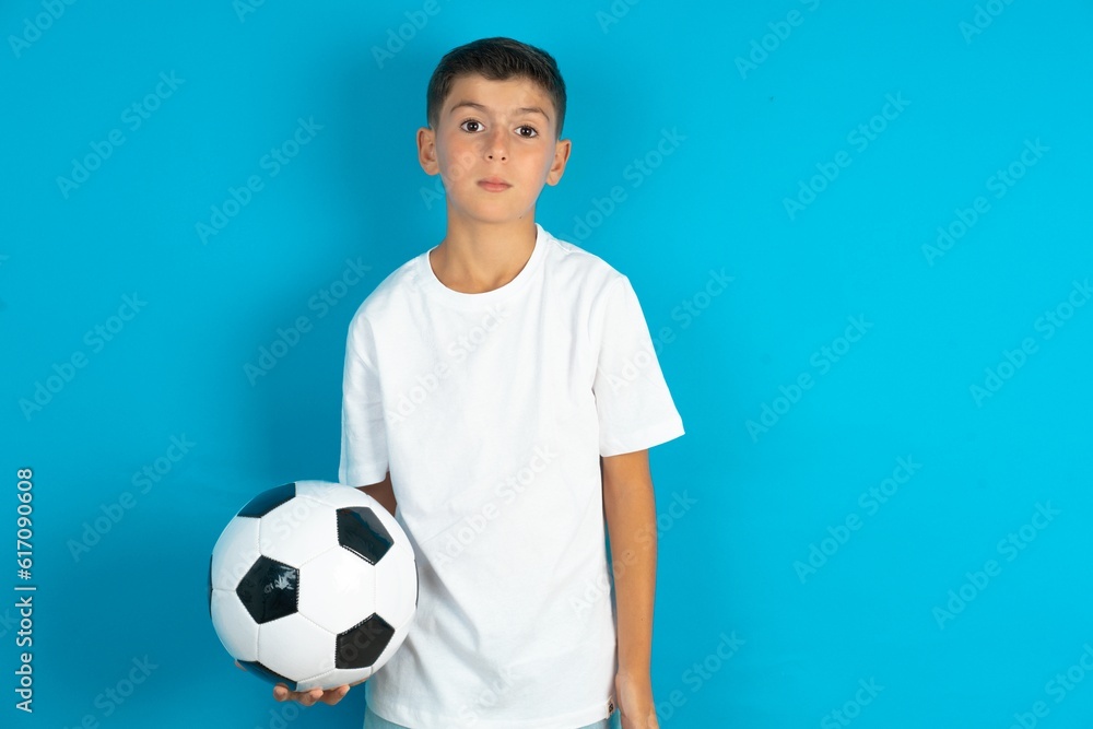 Oh my God. Surprised Little hispanic boy wearing white T-shirt holding a football ball stares at camera with shocked expression exclaims with unexpectedness,