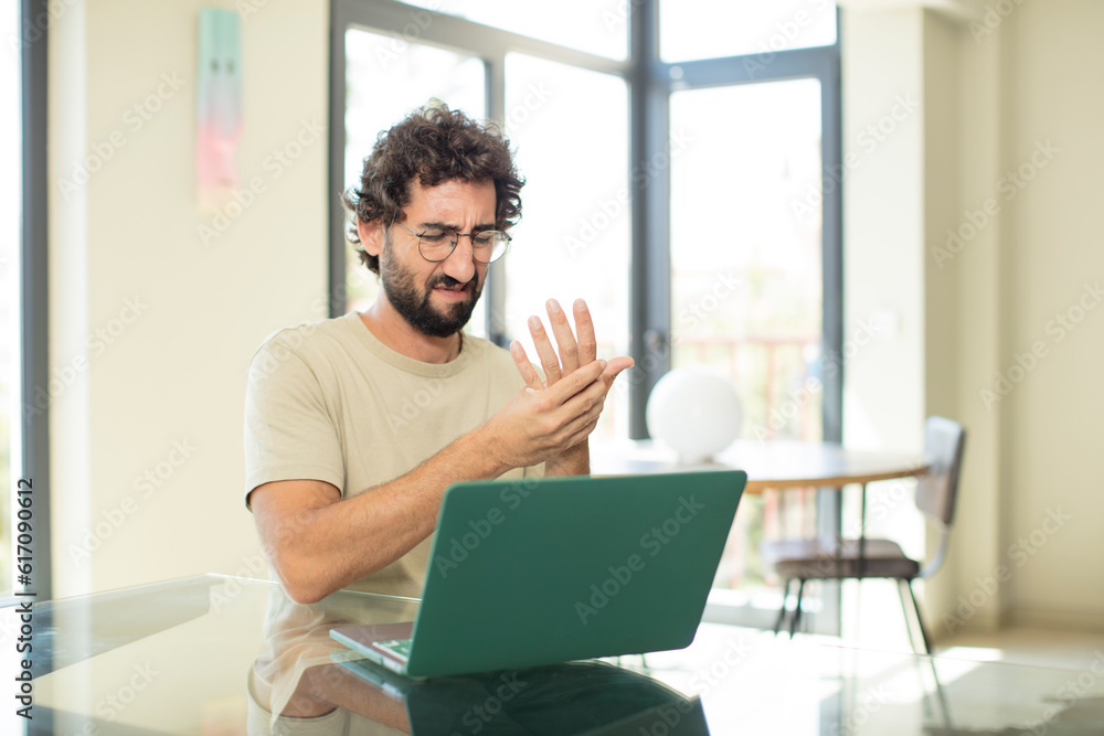 young adult bearded man with a laptop feeling anxious, sick and unhappy, suffering a painful hand ache young adult bearded man with a laptop feeling anxious, sick and unhappy, suffering a painful hand ache