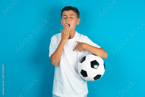 Shocked Little hispanic boy wearing white T-shirt holding a football ball look surprisedly down, indicates at blank with fore finger, Scared model