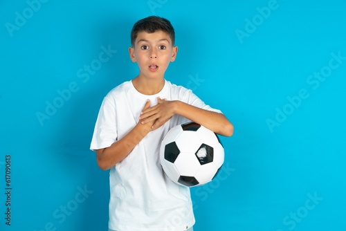 Scared Little hispanic boy wearing white T-shirt holding a football ball looks with frightened expression, keeps hands on chest, being puzzled to notice something strange, People, hush reaction © Jihan