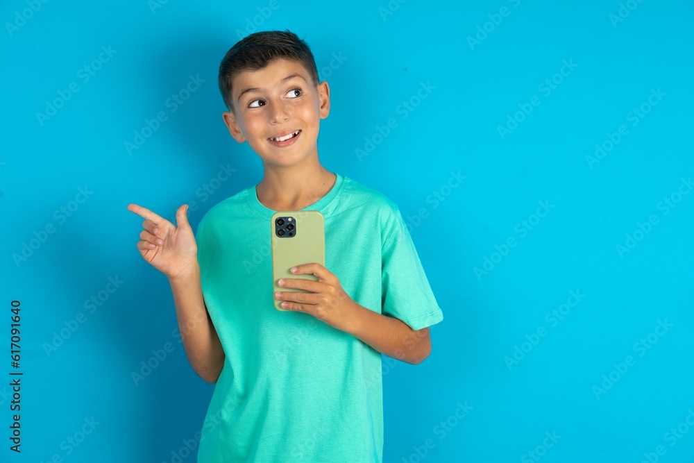 Smiling Little hispanic boy wearing green T-shirt holding mobile phone and pointing aside at copyspace. Smiling Little hispanic boy wearing green T-shirt holding mobile phone and pointing aside at copyspace.