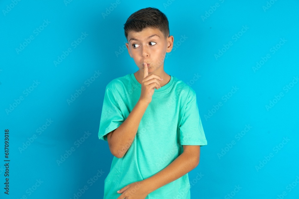 Little hispanic boy wearing green T-shirt silence gesture keeps index finger to lips makes hush sign. Asks not to share secret Little hispanic boy wearing green T-shirt silence gesture keeps index finger to lips makes hush sign. Asks not to share secret