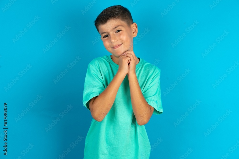 Charming serious Little hispanic boy wearing green T-shirt keeps hands near face smiles tenderly at camera Charming serious Little hispanic boy wearing green T-shirt keeps hands near face smiles tenderly at camera