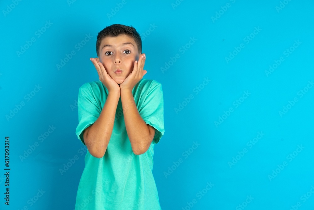 Little hispanic boy wearing green T-shirt with surprised expression keeps hands under chin keeps lips folded makes funny grimace
