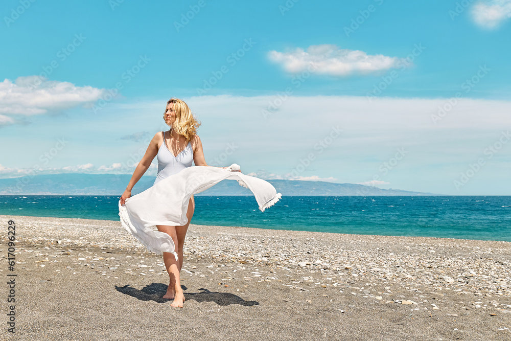 Pretty blond woman in swimsuit and white pareo enjoying sunny windy day near blue sea. Happy female enjoying the life. Beach summer, travel concept.
