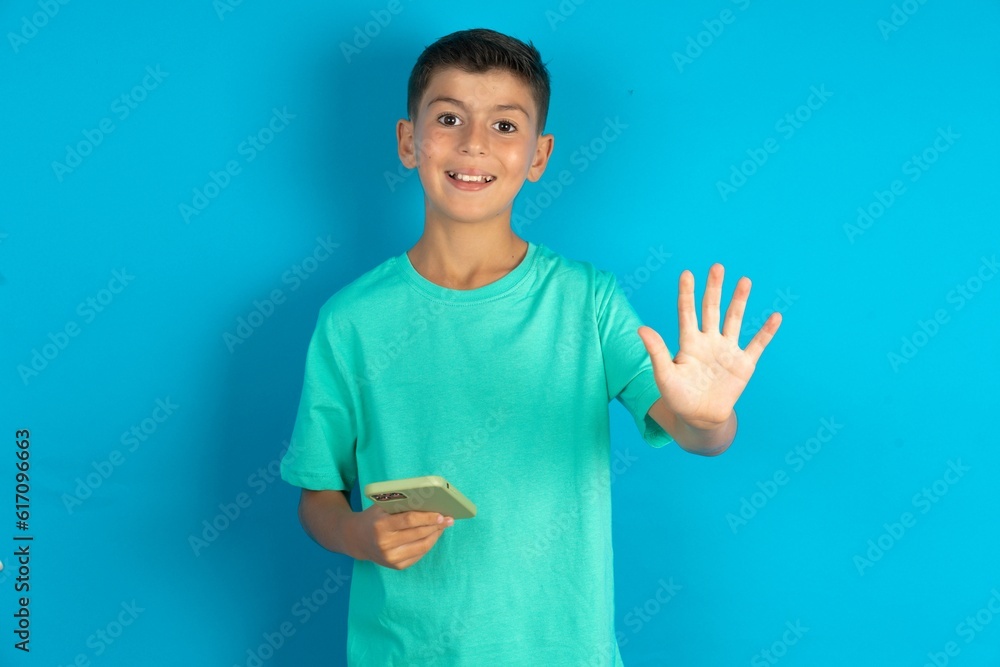 Little hispanic boy wearing green T-shirt using and texting with smartphone with open hand doing stop sign with serious and confident expression, defense gesture Little hispanic boy wearing green T-shirt using and texting with smartphone with open hand doing stop sign with serious and confident expression, defense gesture