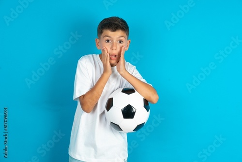 Scared terrified Little hispanic boy wearing white T-shirt holding a soccer ball shocked with prices at shop, People and human emotions concept © Roquillo