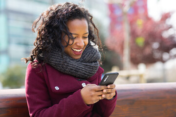Happy young woman in winter wear sitting outside and using mobile phone