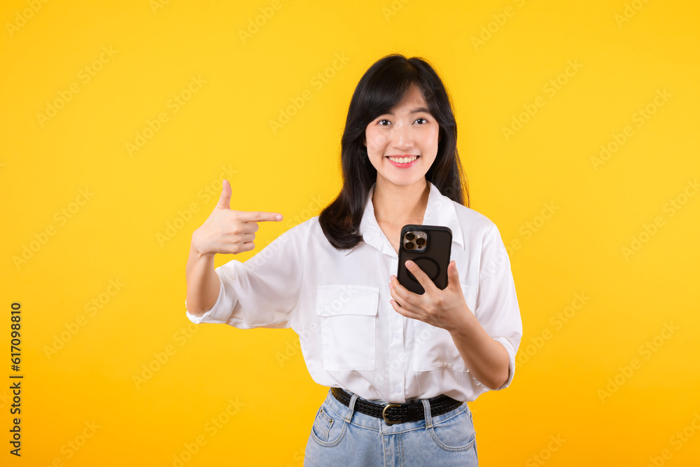Portrait beautiful young asian woman happy smile wearing white shirt and denim plants showing pointing finger hand gesture to smartphone isolated on yellow studio background. app smartphone concept. Portrait beautiful young asian woman happy smile wearing white shirt and denim plants showing pointing finger hand gesture to smartphone isolated on yellow studio background. app smartphone concept.