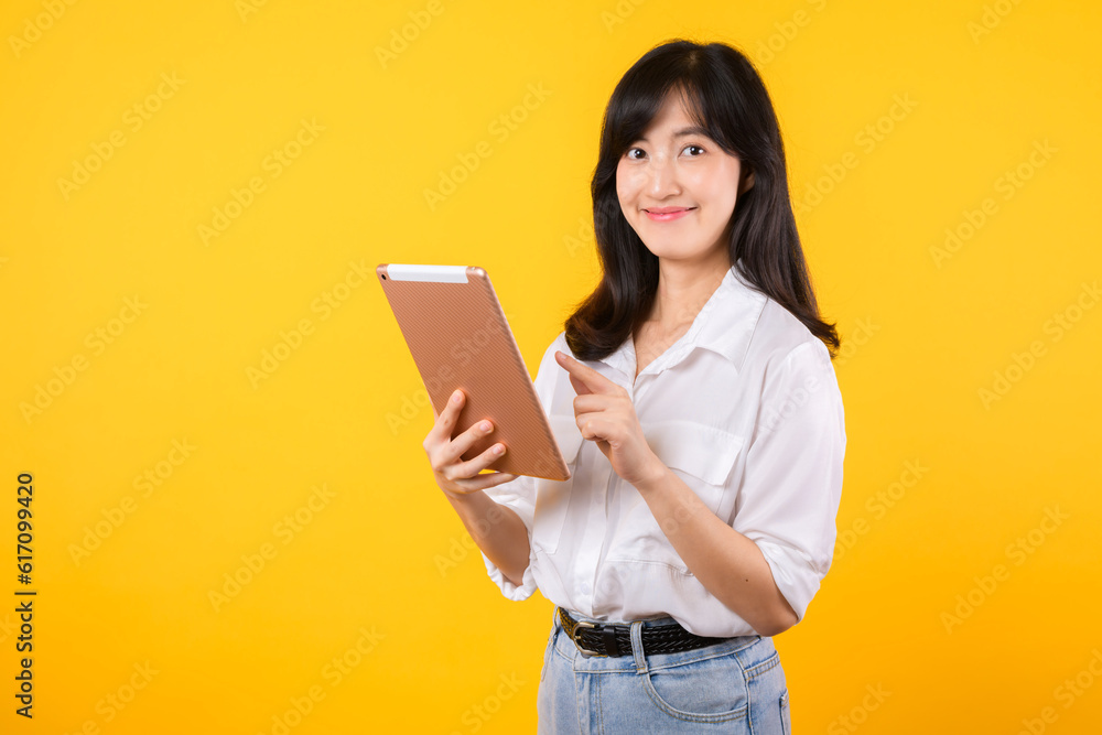 young Asian woman wearing white shirt smiles brightly while holding digital tablet against yellow background, represents a confident company worker embracing modern technology concepts. young Asian woman wearing white shirt smiles brightly while holding digital tablet against yellow background, represents a confident company worker embracing modern technology concepts.