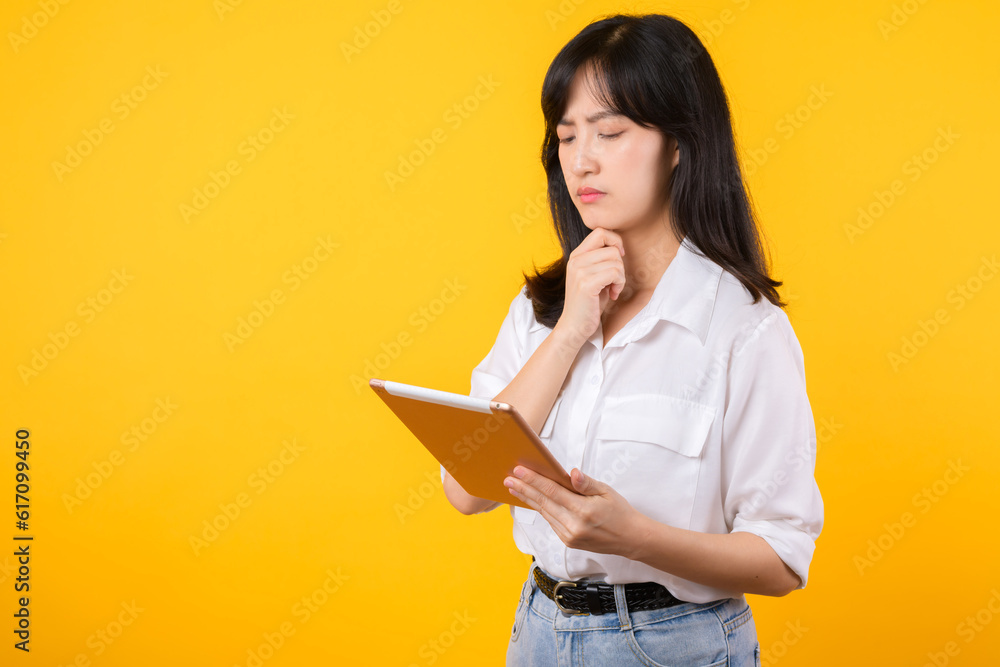 Doubtful Asian young adult business woman working on digital tablet and looking away thinking of solution isolated on yellow background, considering offer, making decision or feeling doubt concept. Doubtful Asian young adult business woman working on digital tablet and looking away thinking of solution isolated on yellow background, considering offer, making decision or feeling doubt concept.