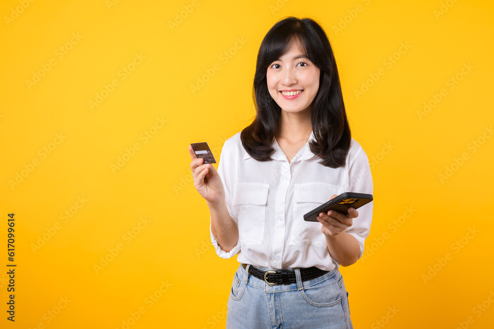 Portrait asian young woman wearing white shirt and denim jean with happy smile holding credit card to paying shopping online on smartphone isolated on yellow background. shopping online concept. Portrait asian young woman wearing white shirt and denim jean with happy smile holding credit card to paying shopping online on smartphone isolated on yellow background. shopping online concept.