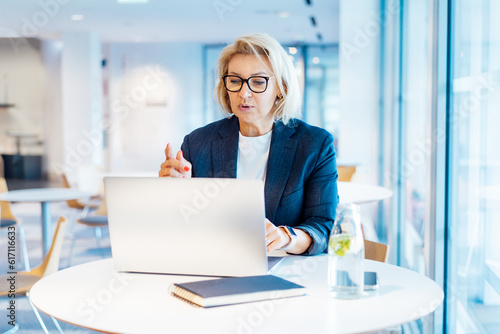 50's confident mature businesswoman having a video call chat on laptop. Middle-aged experienced senior female professional working on laptop in open space office. Female entrepreneur working remotely