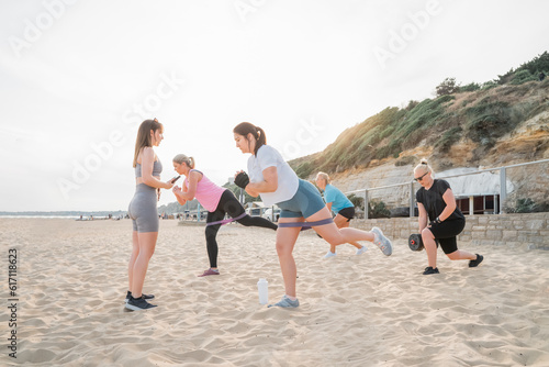 Active women of various ages doing fitness workouts in class exercise with coach on the beach. Ladies working with dumbbells and resistance bands. Sport for health and wellbeing. Active lifestyle