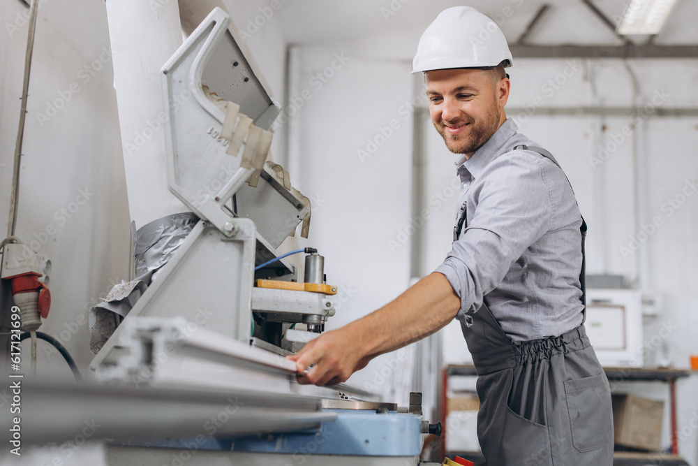 A male worker in a special uniform and an white helmet works on a CNC machine for the production of PVC windows and doors at the factory. A male worker in a special uniform and an white helmet works on a CNC machine for the production of PVC windows and doors at the factory.