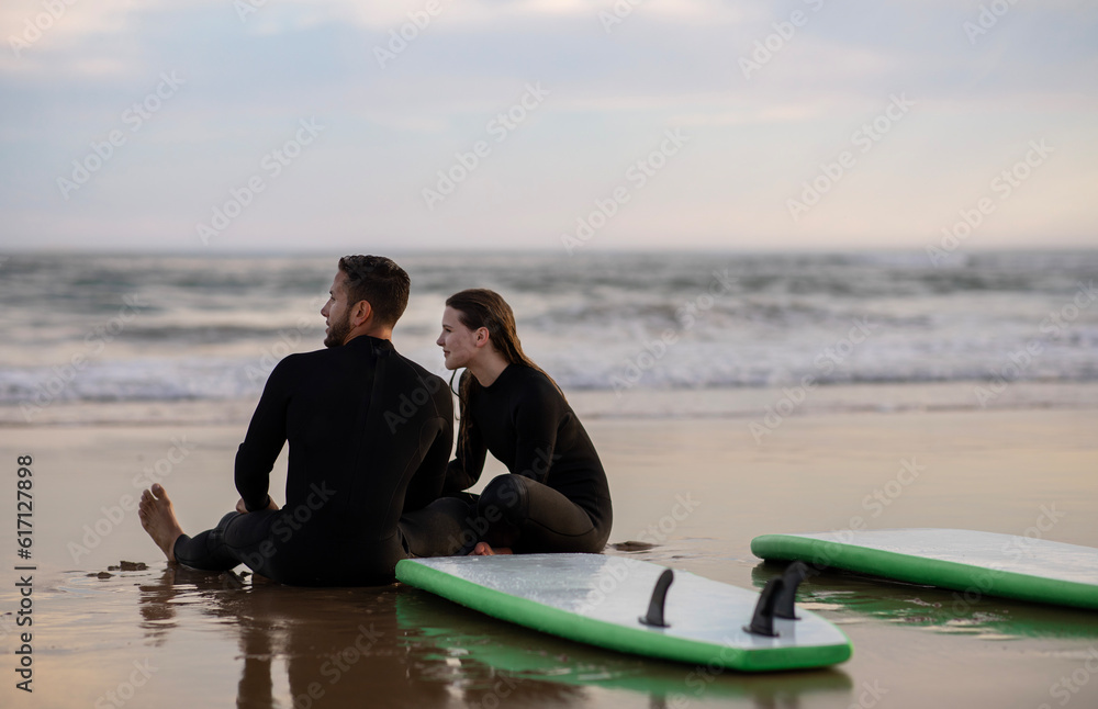 Young Couple In Wetsuits Relaxing On The Beach After Surfing