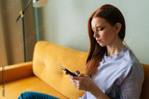 Sad red-haired young woman looking at smartphone screen expression face thinking about problem sitting on yellow couch, difficulty, feeling failure and exhausted, suffering grief and bad relationship