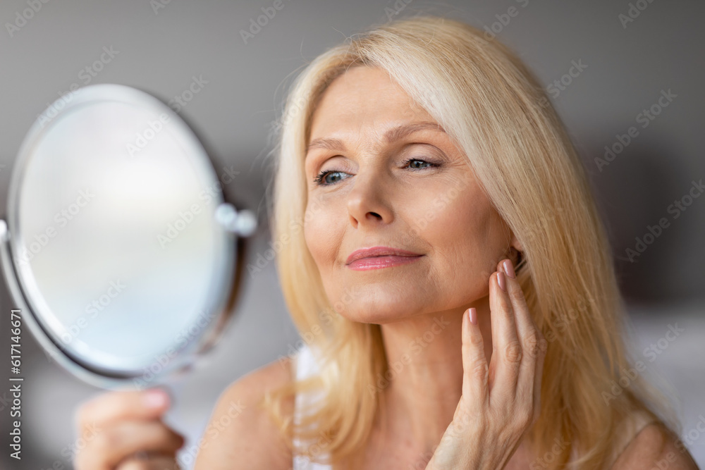 Attractive aged european woman looking in magnifying mirror and touching her face smooth skin, enjoying her appearance