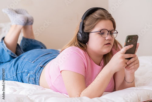 Concentrated overweight teenage girl in headphones lies on bed watching TikTok videos schoolgirl in glasses relaxing at home enjoys social media on smartphone