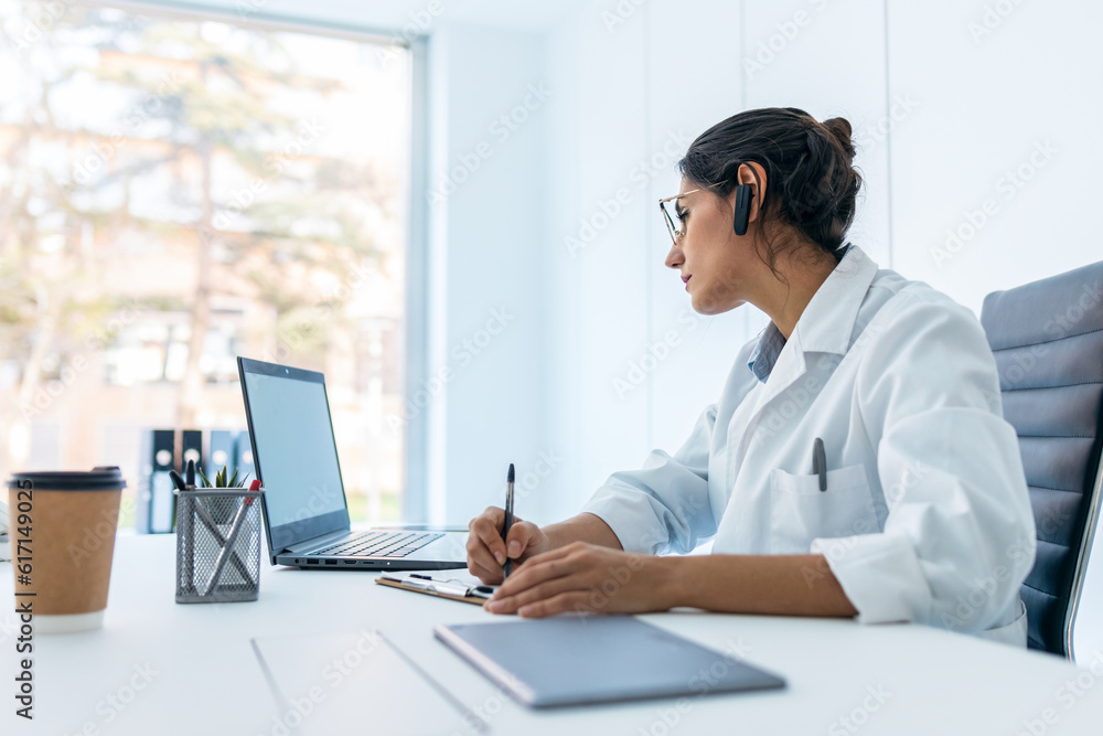 Beautiful female doctor doing a video call while talking with earphone in the medical consultation. Beautiful female doctor doing a video call while talking with earphone in the medical consultation.