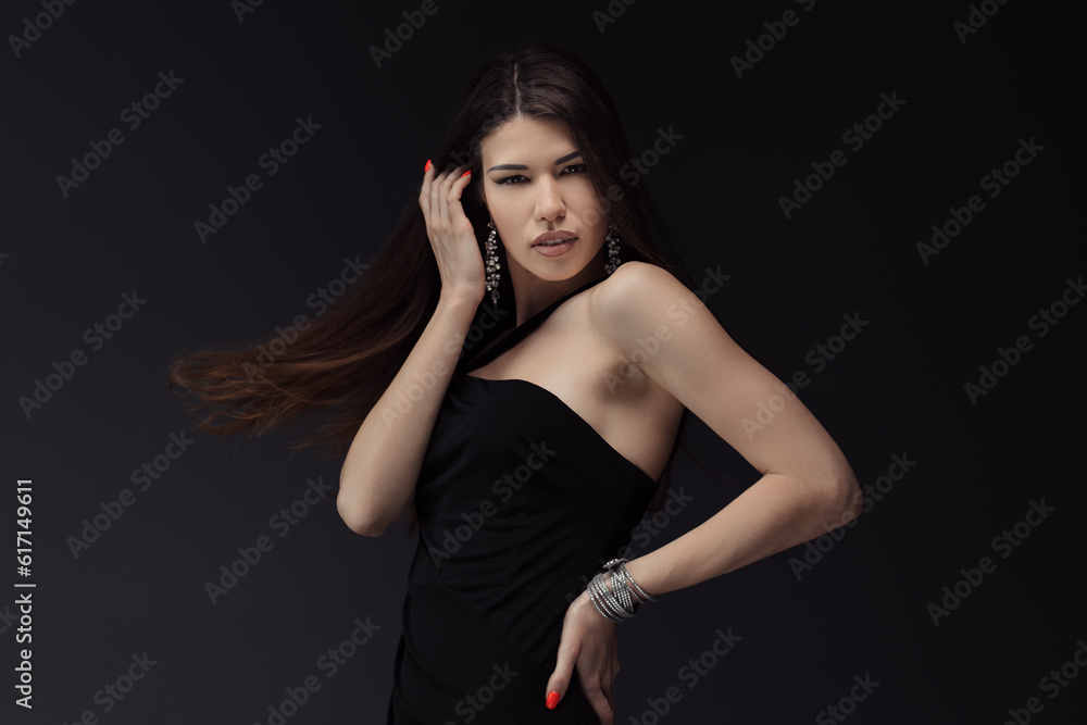 Young attractive female in black dress touching hair and posing for photo session against dark background looking at camera with toothy open mouth Young attractive female in black dress touching hair and posing for photo session against dark background looking at camera with toothy open mouth