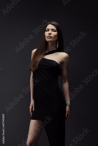 Sensual young female in stylish black dress standing in studio and looking at camera on black background during photo shoot against dark backdrop