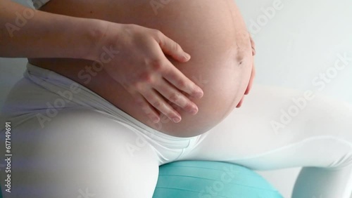 Midsection of heavily pregnant baby bump held by hands of woman bouncing on exercise ball. Side view. White background. Bright shot.
