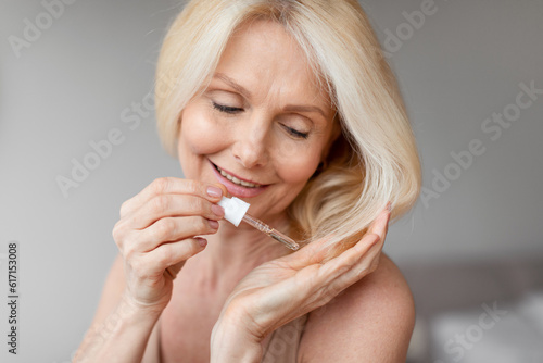 Haircare cosmetics. Happy aged woman applying serum on hair with dropper, sitting in bedroom, free space, closeup