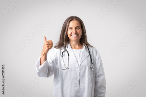 Excited senior female therapist in white coat showing thumb up and smiling  advicing medicine  posing on gray background