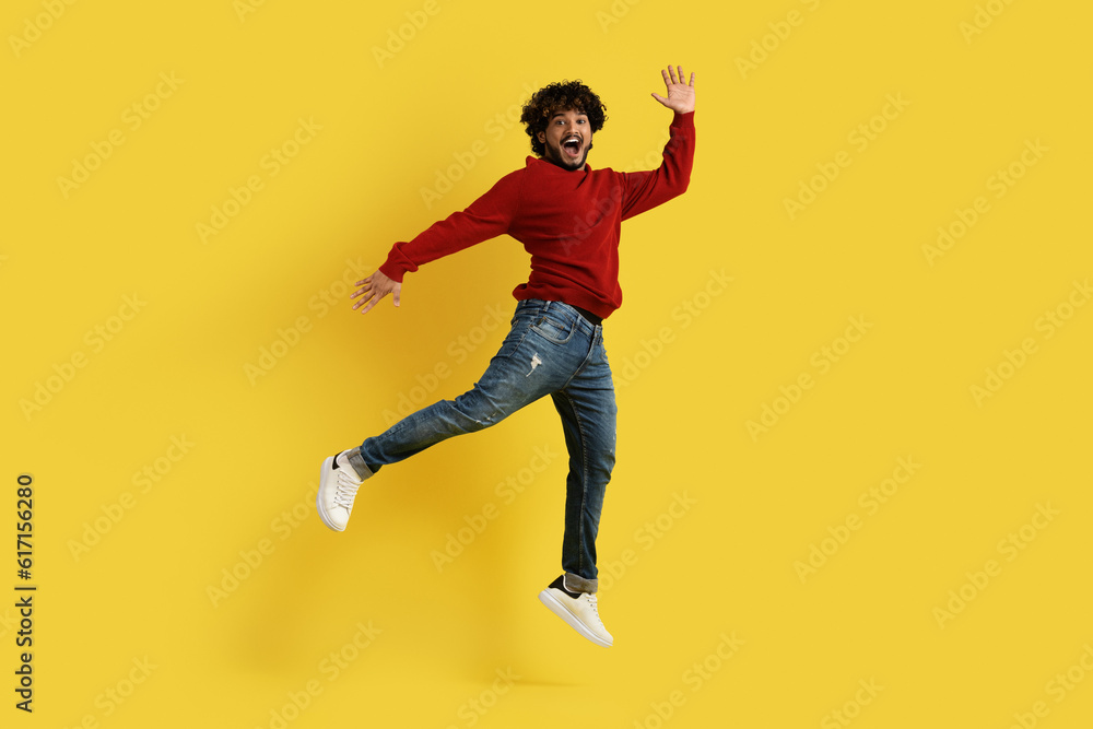 Excited thrilled millennial indian guy jumping over yellow background