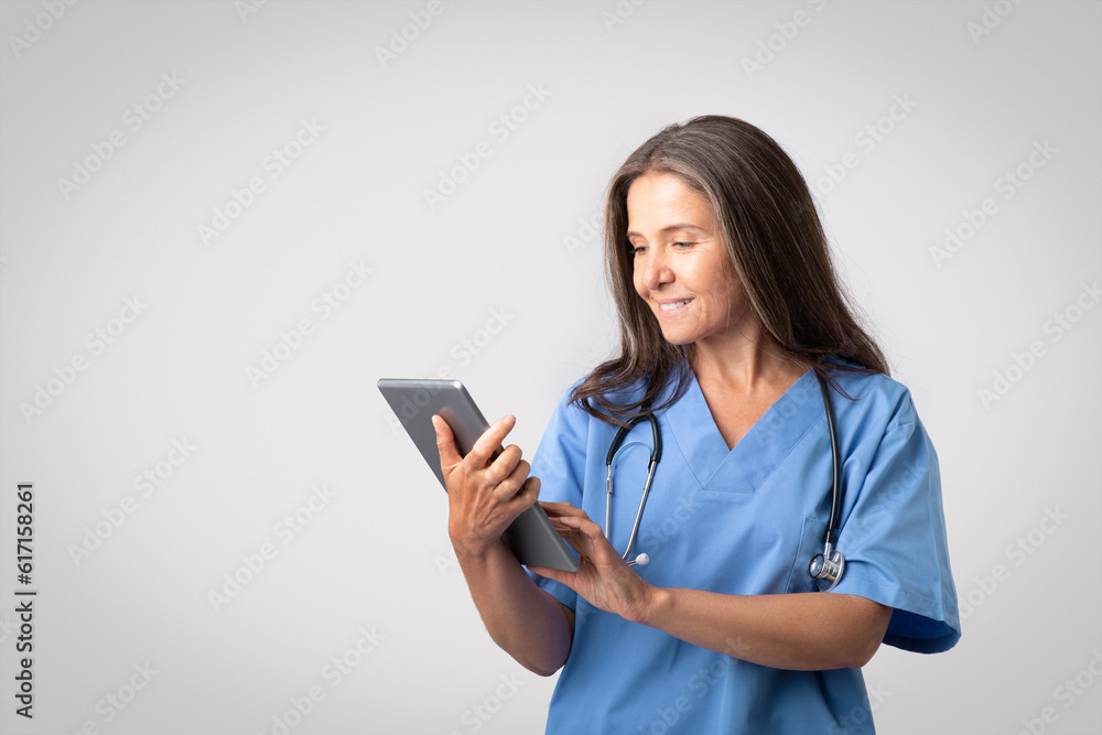 Happy senior european woman doctor in blue uniform typing on digital tablet, posing on light gray background, free space Happy senior european woman doctor in blue uniform typing on digital tablet, posing on light gray background, free space