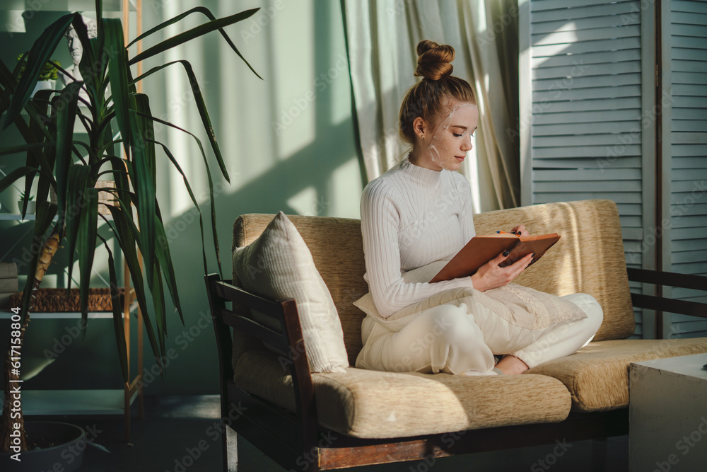Side view of young female freelancer in casual clothes sitting on sofa and reading notes in notebook while working on remote project Side view of young female freelancer in casual clothes sitting on sofa and reading notes in notebook while working on remote project