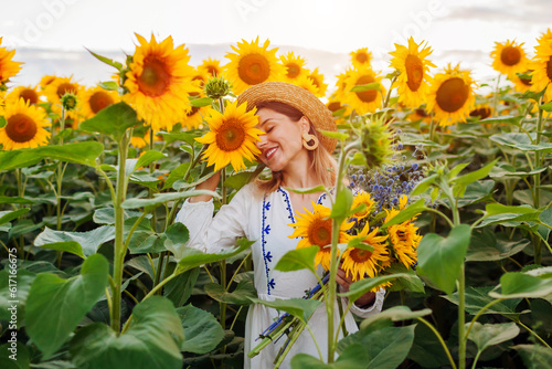 Portrait of young woman walking in blooming sunflower field at sunset smelling picking flowers. Summer in Ukraine