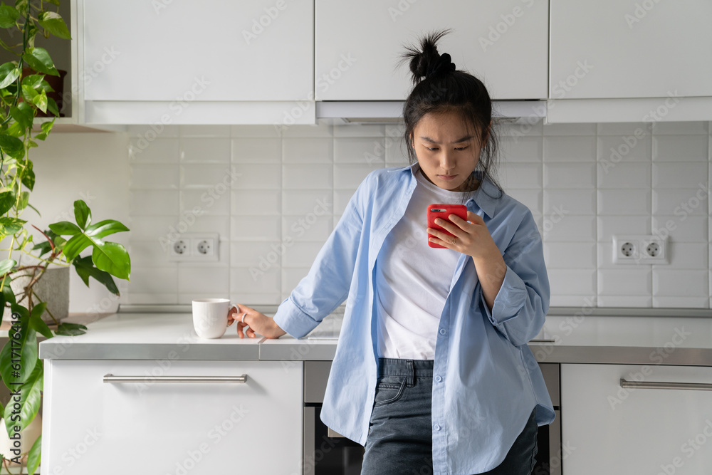 Φωτογραφία Upset young Asian woman standing in kitchen holding mobile phone, reading unpleasant sms on cellphone, receiving message with bad news