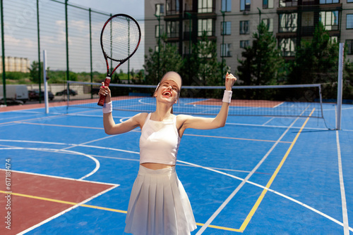 young girl tennis player in white uniform with racket rejoices in victory and shouts on blue court outdoors © Богдан Маліцький