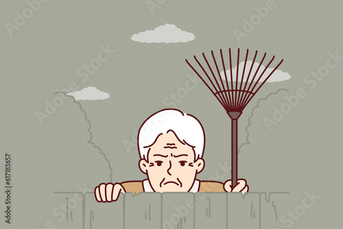 Elderly envious man neighbor peeps from behind fence spying on residents of private house. Envious male pensioner with rake looking at screen suspiciously for bad neighborhood concept.