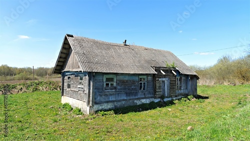 On a grassy lawn stands an old wooden house with boarded-up windows and a slate roof. Next to it are trees and shrubs. Behind the house is a farm field and a road, and behind it is a forest. Sunny