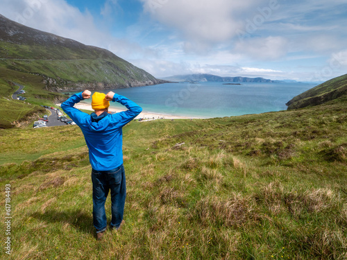 Male tourist in blue jacket and yellow hat looking at stunning nature scene with beach, ocean, mountain and cliffs. Warm sunny day. Keem bay, Ireland. Travel and tourism. Irish nature landscape.