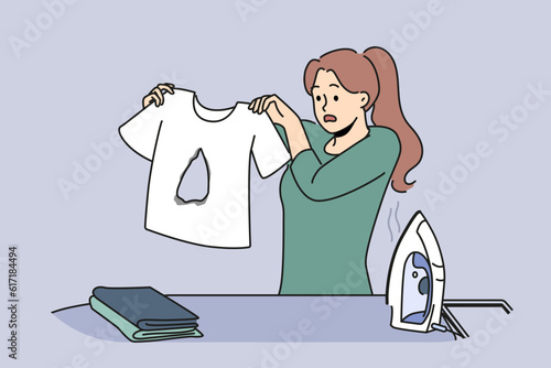 Clumsy housewife ironing clothes burnt hole in t-shirt and dont know how to fix mistake. Woman stands near ironing board with clothes and iron and looks at damaged item in confusion.
