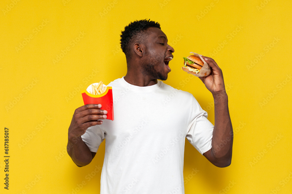 happy african american man in white t-shirt holding big burger and french fries and smiling, guy eating fast food happy african american man in white t-shirt holding big burger and french fries and smiling, guy eating fast food