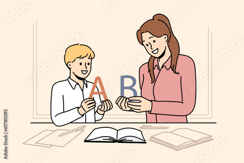 Back to school concept with boy learning alphabet along with elementary school teacher standing near table with books. Woman teaches child read or spell correctly, wanting give son quality education