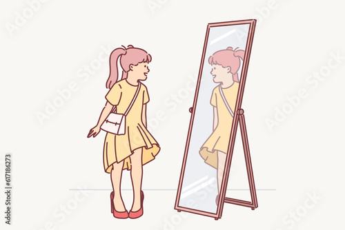 Little girl in mom clothes looks in mirror trying on shoes with heel and trying to seem like adult