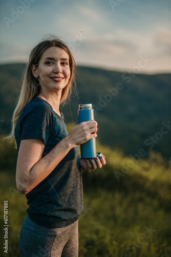 Fitness motivation: Depict a woman drinking water in a sports outfit amidst breathtaking natural surroundings, inspiring viewers to engage in physical activities and prioritize their well-being.