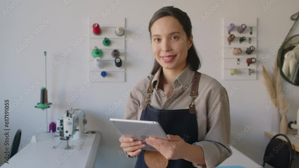 Close-up tilting portrait shot of female fashion designer standing at cutting table in atelier with pieces of leather and product drawings, using tablet computer, looking at camera and smiling Close-up tilting portrait shot of female fashion designer standing at cutting table in atelier with pieces of leather and product drawings, using tablet computer, looking at camera and smiling