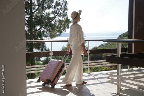Tourist woman with sun hat carrying wheeled luggage walking in resort loking for her room. Travelling, vacation, leisure, summer holiday, solo traveling concept.
