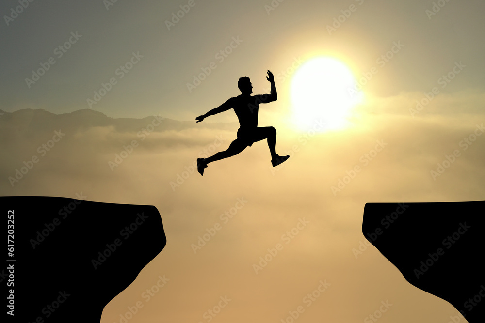 Concept of reaching life and business goals. Silhouette of man jumping over chasm at sunrise