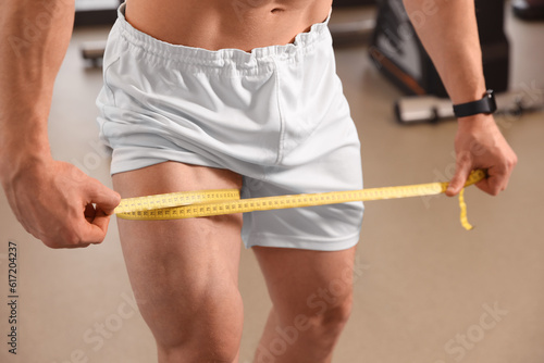 Athletic man measuring thigh with tape in gym, closeup
