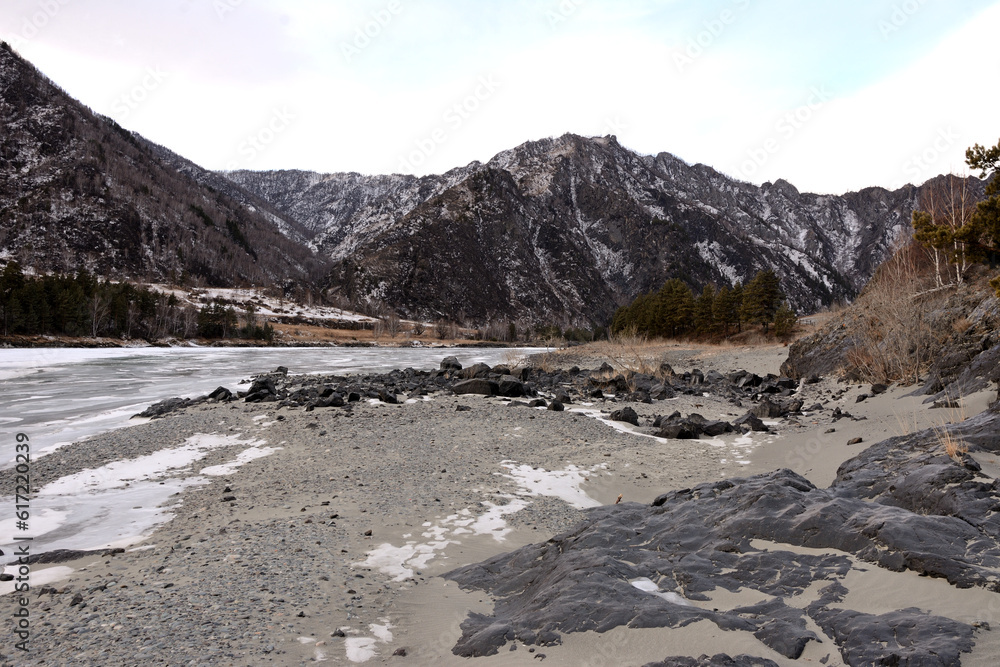 Protruding flat stones on the sandy bank of a frozen river flowing through the valley on a sunny winter evening.